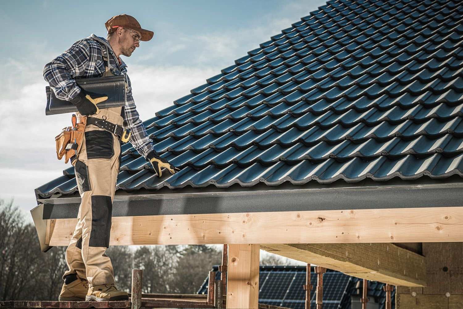 A roofer on scaffolding checks dark interlocking tiles on a newly built roof, with solar panels on a roof behind