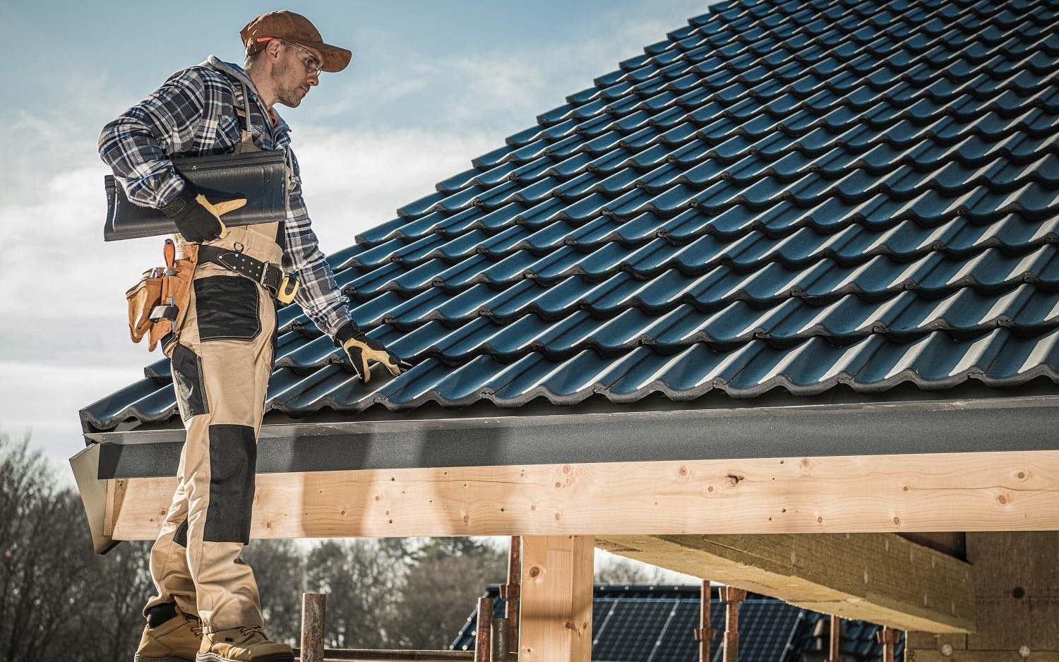 A roofer on scaffolding checks dark interlocking tiles on a newly built roof, with solar panels on a roof behind