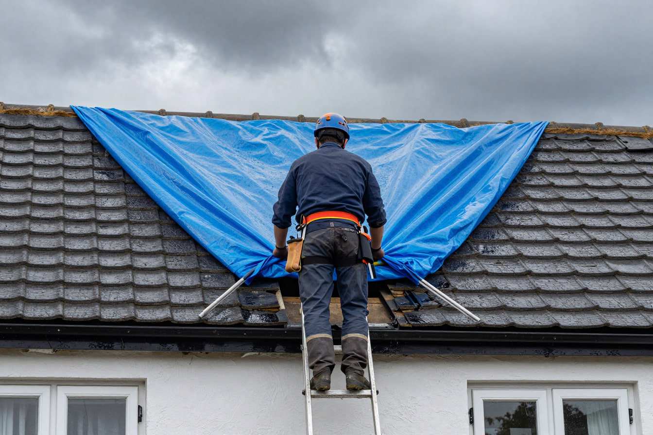 Roofer securing a temporary blue tarpaulin over a storm-damaged section of a tiled roof